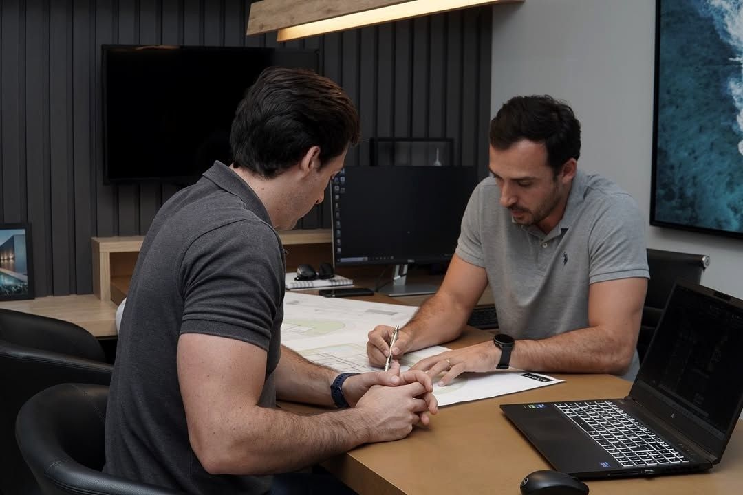 Fotografia de dois profissionais do escritório debruçados sobre uma prancha impressa de projeto, em sala com parede ripada escura.