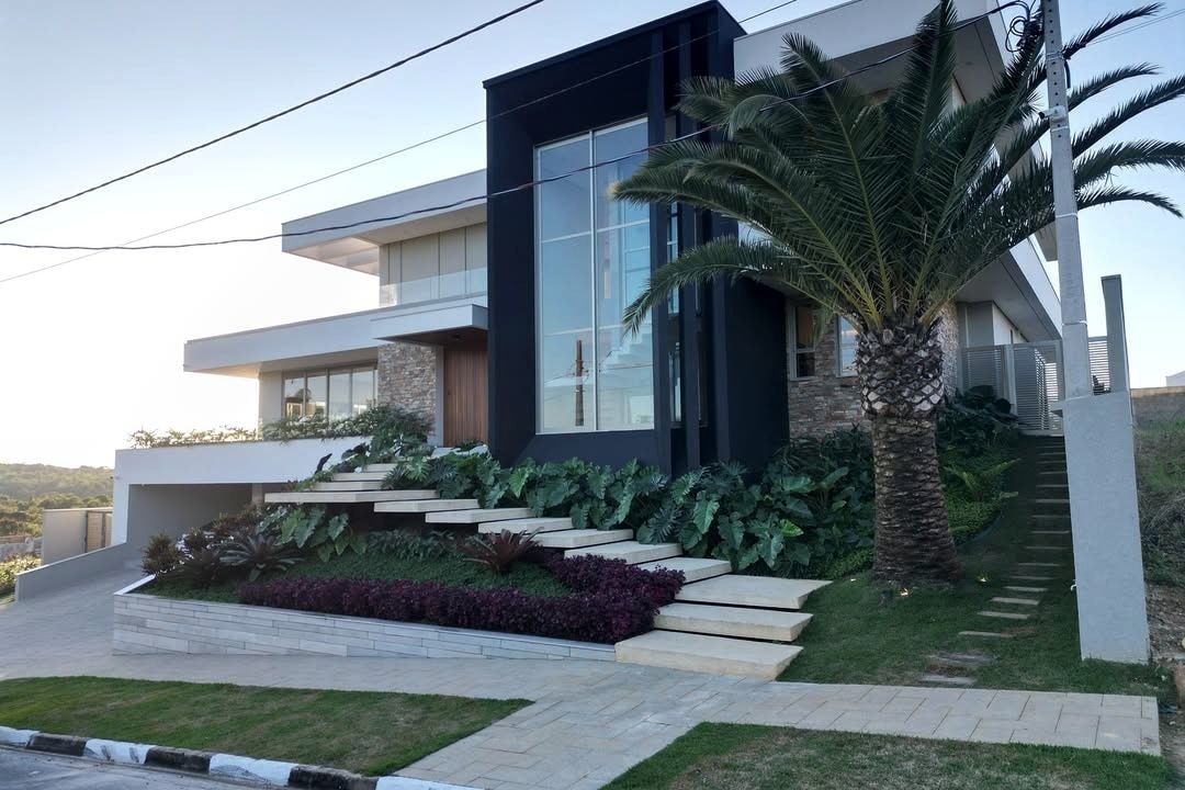 Fotografia da mesma casa já construída, vista da rua ao fim da tarde: o mesmo volume envidraçado escuro, a mesma palmeira já crescida, os degraus de pedra em meio ao paisagismo, os morros ao fundo e os fios de energia cruzando o céu.