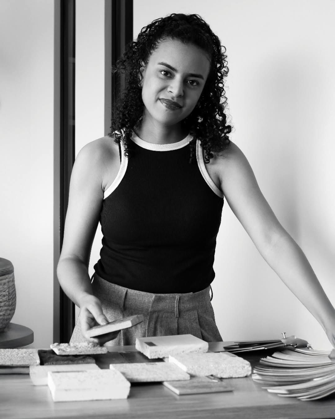 Retrato em preto e branco de Taiane, arquiteta e urbanista do escritório, diante de amostras de revestimento sobre a mesa.
