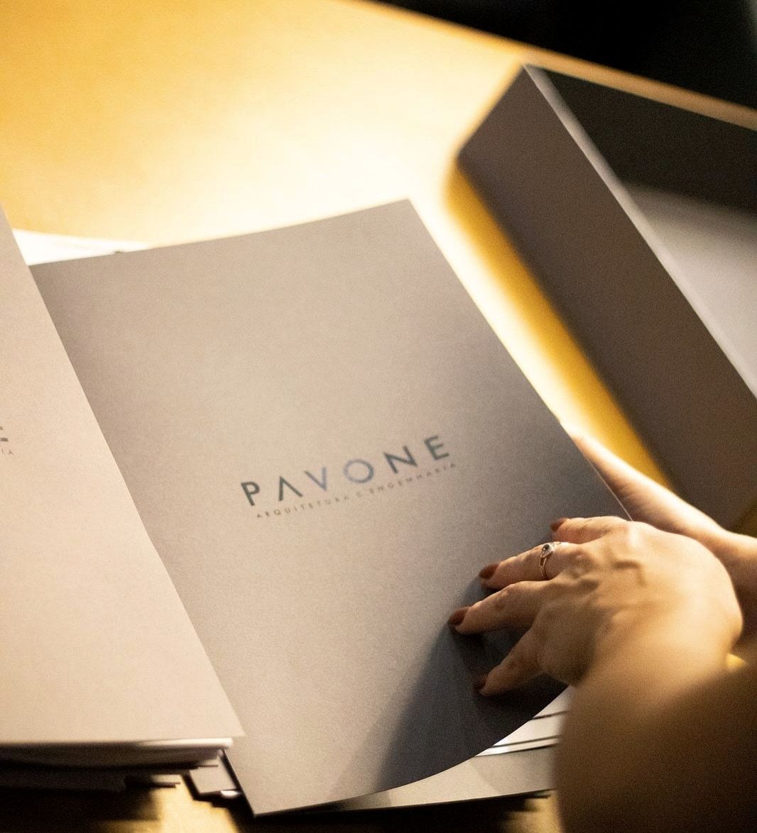 Fotografia de mãos abrindo a pasta impressa com o logotipo Pavone Arquitetura e Engenharia sobre a mesa de trabalho.