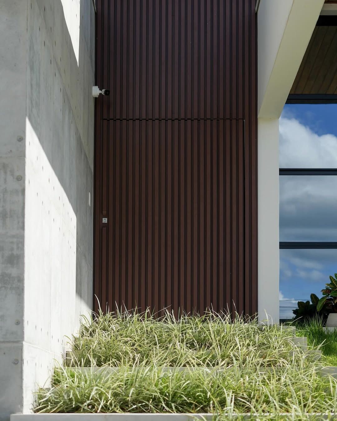 Fotografia da obra da Casa AF: porta pivotante ripada entre concreto aparente e canteiro.